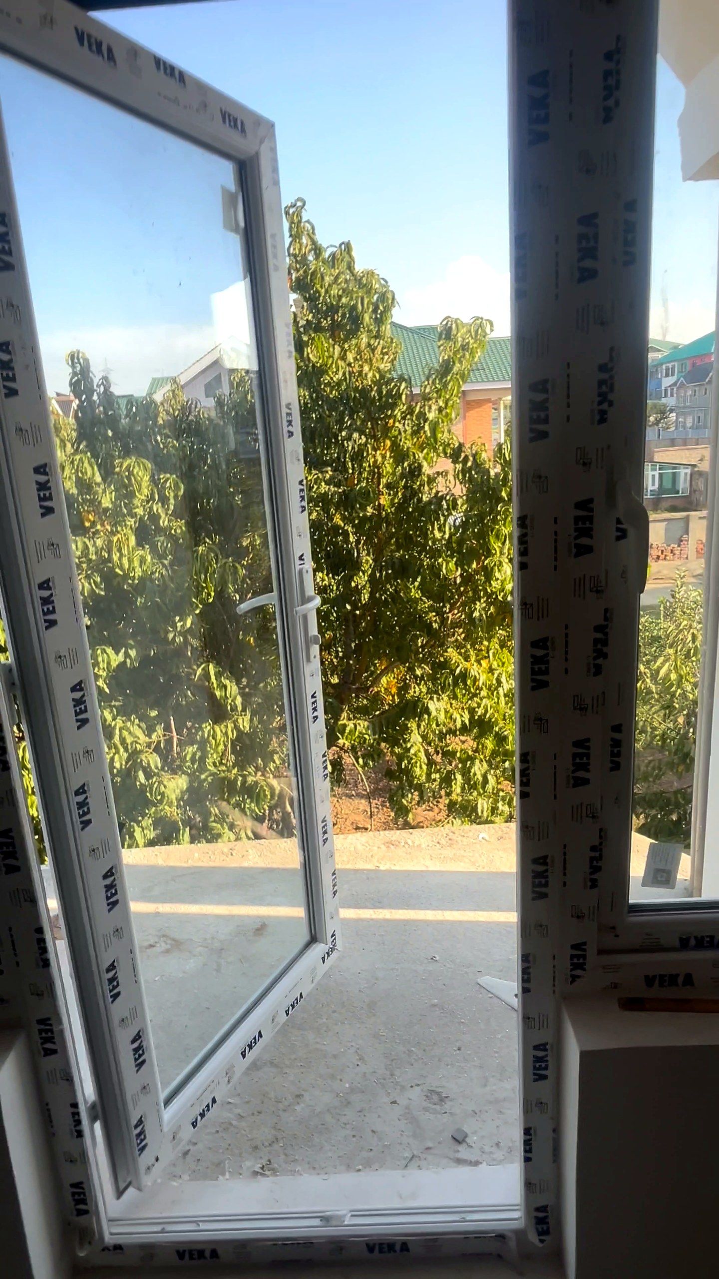 Casement window opened onto trees, with the VEKA protective film still on the frame